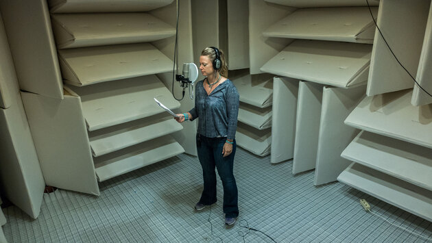 woman standing in middle of a room with walls covered with insulated acoustic wedges. She wears headphones and are talking into a midronphone.