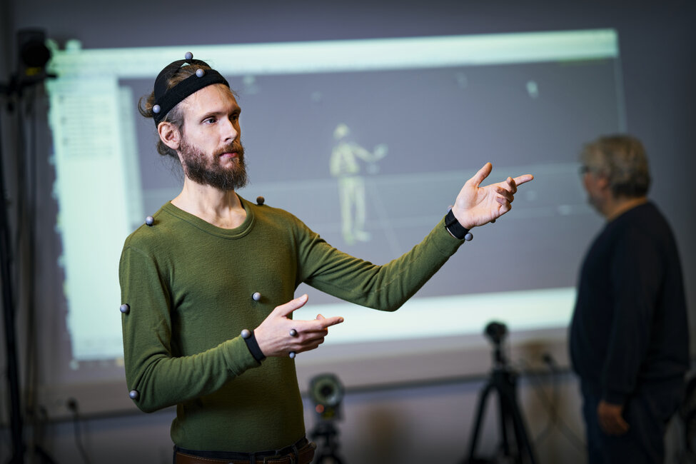 man with small reflektive round markers on his head and upper body stands in fron of a screen where a visualisation of markers are shown