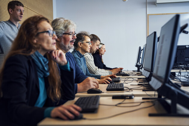 a row of people are sitting and looking at screens with eye trackers. One man is standing behind them.