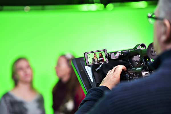 two persons in front of a green screen being filmed by another person