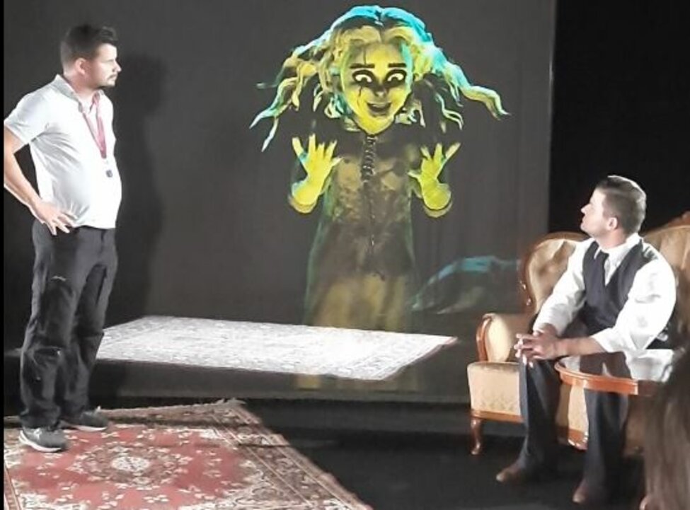 A theater scene with two actors looking at a avatar on a screen between them