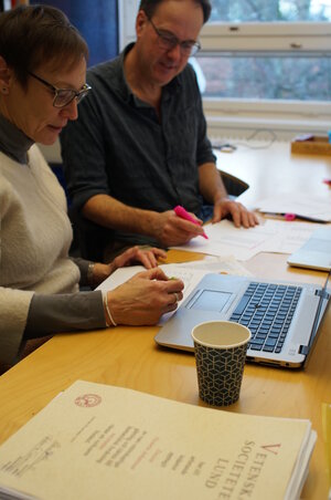 a man and a woman sitting at a desk with computers and papers on it. They are discussing something.