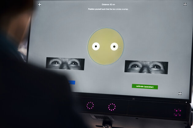 i kanten på bilden syns en axel bakifrån på en person som tittar på en skärm. på skärmen syns en rund figur samt en inspelning på personens ögon.