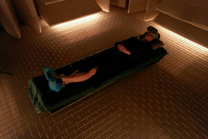 [Translate to Svenska:] woman lying on a mattress on a mesh floor. She is wearing a VR-headset and soft light comes from below the walls that is made of a sound insulated material.