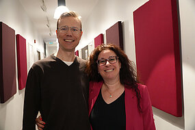 portrait picture, blond man with glasses and brown sweater next to woman with dark, curly hair and glasses, red cardigan. Both are smiling.