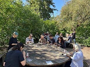 [Translate to English:] people sitting around a big round table outside having lunch