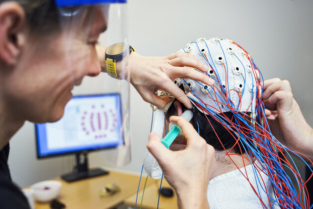 one person is sitting with a cap with sensors and cords on the head, looking at a screen with data, while another person is applying gel to the sensors with a syringe.