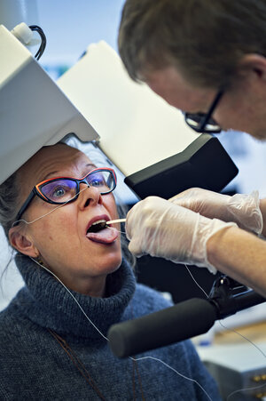 kvinna sitter under vita maskinarmar. En man med handskar sätter sensorer på hennes tunga.