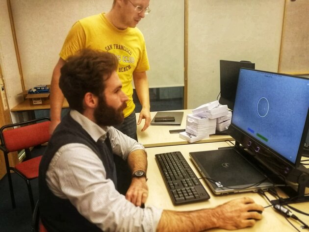 Two men, one sitting and one standing, in front of computer with eyetracker. 